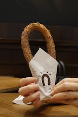 A hand is holding a churro wrapped in a paper sleeve with a stylized design that resembles an arch or horseshoe. The background is a wooden table and dimly lit interior, suggesting a cozy setting.