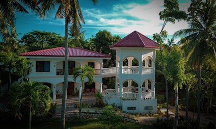 A luxurious two-story villa with a red-tiled roof surrounded by lush tropical greenery and tall palm trees. The building features white arches and a spacious balcony. A manicured garden and a cobblestone path enhance the scenery.