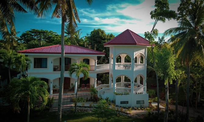 A luxurious two-story villa with a red-tiled roof surrounded by lush tropical greenery and tall palm trees. The building features white arches and a spacious balcony. A manicured garden and a cobblestone path enhance the scenery.
