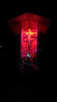 A crucifix is illuminated against a dark background with vibrant red lighting. The structure is adorned with colorful, glowing lights, enhancing its visual impact. A roof covers the scene, casting a dramatic and spiritual atmosphere.