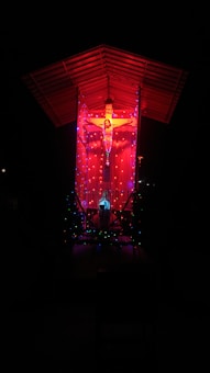 A crucifix is illuminated against a dark background with vibrant red lighting. The structure is adorned with colorful, glowing lights, enhancing its visual impact. A roof covers the scene, casting a dramatic and spiritual atmosphere.
