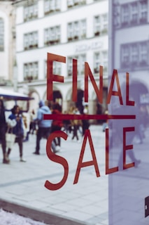 Large red letters spelling 'FINAL SALE' are displayed on a glass window. In the background, a busy pedestrian area can be seen with people walking and historic buildings in view, slightly blurred, creating a sense of movement and activity.