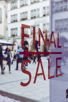 Large red letters spelling 'FINAL SALE' are displayed on a glass window. In the background, a busy pedestrian area can be seen with people walking and historic buildings in view, slightly blurred, creating a sense of movement and activity.