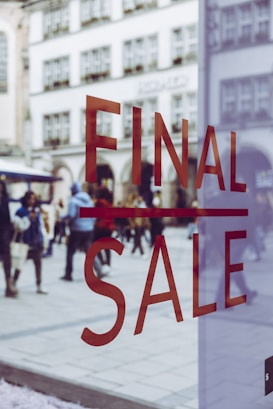 Large red letters spelling 'FINAL SALE' are displayed on a glass window. In the background, a busy pedestrian area can be seen with people walking and historic buildings in view, slightly blurred, creating a sense of movement and activity.