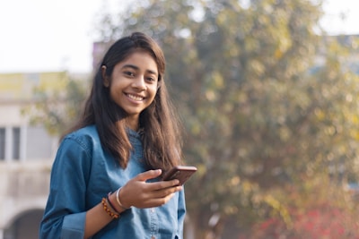 A young woman holding a smartphone standing on a street, smiling confidently.