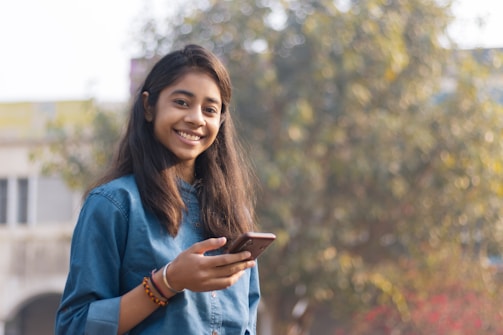 A close-up of a woman smiling while holding her phone outdoors.