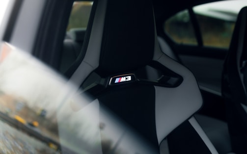 A close-up of a car seat with the 'M3' logo visible, highlighting the interior of a luxury vehicle. The seat is upholstered in a combination of dark and light materials, and the surrounding environment is slightly out of focus.