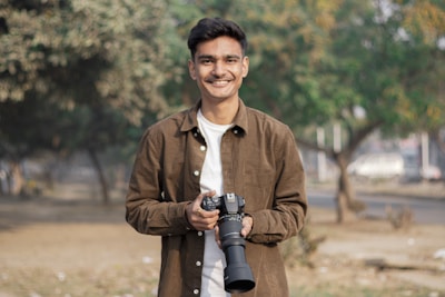 A person stands in an outdoor setting holding a camera with a large lens. The individual is wearing a brown jacket and a white shirt, smiling with a natural background featuring blurred green trees and a pathway.