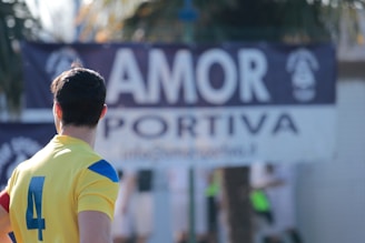 A person wearing a yellow sports jersey with the number 4 stands with their back to the camera. The background features a blurred banner displaying the word 'AMOR'.