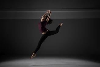 A person is captured in mid-air, performing a dynamic and graceful dance leap against a dark, minimalist background. The individual wears a maroon top and black pants and appears to be barefoot.