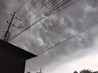 Dark, heavy clouds dominate the sky, signaling an impending storm. Power lines and a utility pole create a network across the scene, with a building's roof partially visible in the lower left corner. The atmosphere is overcast with a looming, dramatic feel.
