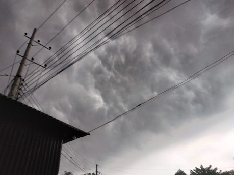 Dark, heavy clouds dominate the sky, signaling an impending storm. Power lines and a utility pole create a network across the scene, with a building's roof partially visible in the lower left corner. The atmosphere is overcast with a looming, dramatic feel.