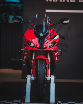 A vibrant red sport motorcycle is displayed on a stand, positioned front and center. The bike features sleek and aerodynamic design elements, including a black windscreen and angular mirrors. The lighting accentuates its sharp lines and modern aesthetic.