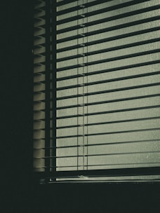 Horizontal blinds covering a window, with soft light filtering through, casting subtle shadows. The scene has a muted and slightly abstract feel, suggesting an indoor environment.