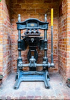 A vintage industrial metal press with a large lever is positioned against a brick wall. The machine features intricate metalwork and a plaque that might indicate its manufacturer. The bricks are light red and orange, giving a rustic background contrast to the dark metal of the press.