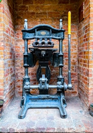 A vintage industrial metal press with a large lever is positioned against a brick wall. The machine features intricate metalwork and a plaque that might indicate its manufacturer. The bricks are light red and orange, giving a rustic background contrast to the dark metal of the press.