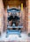 A vintage industrial metal press with a large lever is positioned against a brick wall. The machine features intricate metalwork and a plaque that might indicate its manufacturer. The bricks are light red and orange, giving a rustic background contrast to the dark metal of the press.