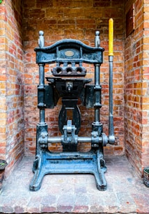 A vintage industrial metal press with a large lever is positioned against a brick wall. The machine features intricate metalwork and a plaque that might indicate its manufacturer. The bricks are light red and orange, giving a rustic background contrast to the dark metal of the press.
