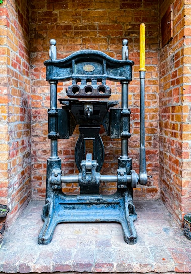 A vintage industrial metal press with a large lever is positioned against a brick wall. The machine features intricate metalwork and a plaque that might indicate its manufacturer. The bricks are light red and orange, giving a rustic background contrast to the dark metal of the press.