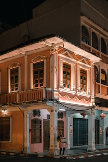 An elegant two-story building with intricate architectural details, featuring ornate decorations and illuminated by warm lighting. The facade is painted in pastel colors with decorative moldings, and a person stands at the corner holding some boxes. Traditional lanterns hang near the entrance, adding an element of cultural ambiance.
