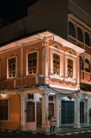An elegant two-story building with intricate architectural details, featuring ornate decorations and illuminated by warm lighting. The facade is painted in pastel colors with decorative moldings, and a person stands at the corner holding some boxes. Traditional lanterns hang near the entrance, adding an element of cultural ambiance.