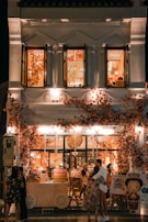 A beautifully decorated shop with an abundance of flowers adorning the facade, creating a warm and inviting atmosphere. The shop is illuminated with soft, ambient lighting, and features people standing and embracing outside. The second floor displays open windows with more floral decorations inside.