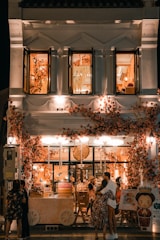 A beautifully decorated shop with an abundance of flowers adorning the facade, creating a warm and inviting atmosphere. The shop is illuminated with soft, ambient lighting, and features people standing and embracing outside. The second floor displays open windows with more floral decorations inside.