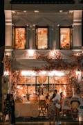 A beautifully decorated shop with an abundance of flowers adorning the facade, creating a warm and inviting atmosphere. The shop is illuminated with soft, ambient lighting, and features people standing and embracing outside. The second floor displays open windows with more floral decorations inside.