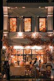 A beautifully decorated shop with an abundance of flowers adorning the facade, creating a warm and inviting atmosphere. The shop is illuminated with soft, ambient lighting, and features people standing and embracing outside. The second floor displays open windows with more floral decorations inside.