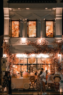 A beautifully decorated shop with an abundance of flowers adorning the facade, creating a warm and inviting atmosphere. The shop is illuminated with soft, ambient lighting, and features people standing and embracing outside. The second floor displays open windows with more floral decorations inside.