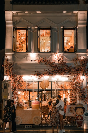A beautifully decorated shop with an abundance of flowers adorning the facade, creating a warm and inviting atmosphere. The shop is illuminated with soft, ambient lighting, and features people standing and embracing outside. The second floor displays open windows with more floral decorations inside.