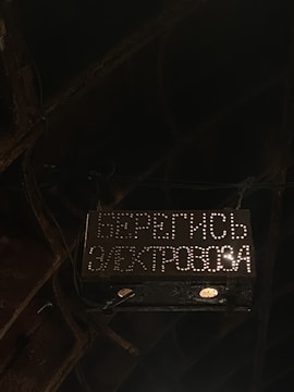 A dark and industrial setting with a warning sign. The sign is illuminated with white dots forming Cyrillic text, warning of an electric hazard. Surrounding the sign are metal structures and wires, creating a gritty atmosphere.