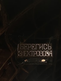 A dark and industrial setting with a warning sign. The sign is illuminated with white dots forming Cyrillic text, warning of an electric hazard. Surrounding the sign are metal structures and wires, creating a gritty atmosphere.