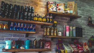 A set of wooden shelves mounted on a brick wall displays various packaged food and beverage products. The assortment includes energy drink cans, coffee packets, and bottles. Some shelves feature brightly colored coffee makers and assorted snacks like chocolate bars.