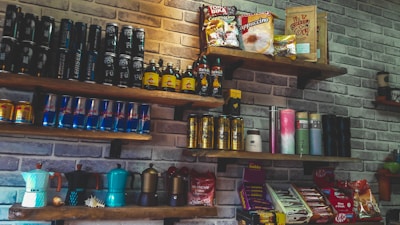 A set of wooden shelves mounted on a brick wall displays various packaged food and beverage products. The assortment includes energy drink cans, coffee packets, and bottles. Some shelves feature brightly colored coffee makers and assorted snacks like chocolate bars.