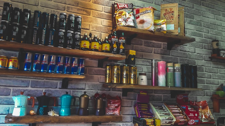 A set of wooden shelves mounted on a brick wall displays various packaged food and beverage products. The assortment includes energy drink cans, coffee packets, and bottles. Some shelves feature brightly colored coffee makers and assorted snacks like chocolate bars.