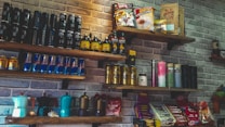 A set of wooden shelves mounted on a brick wall displays various packaged food and beverage products. The assortment includes energy drink cans, coffee packets, and bottles. Some shelves feature brightly colored coffee makers and assorted snacks like chocolate bars.