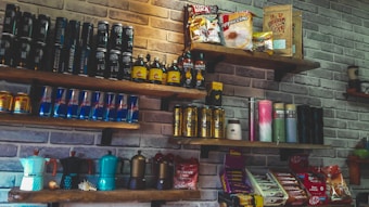 A set of wooden shelves mounted on a brick wall displays various packaged food and beverage products. The assortment includes energy drink cans, coffee packets, and bottles. Some shelves feature brightly colored coffee makers and assorted snacks like chocolate bars.