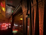 The interior of a bus at night is dimly lit with warm, ambient lighting. Heavy curtains cover the window, and reflections of red taillights from traffic outside can be seen through the glass. Air conditioning vents are visible on the bus ceiling, and a small panel or sign is positioned near the window.
