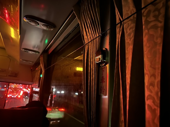 The interior of a bus at night is dimly lit with warm, ambient lighting. Heavy curtains cover the window, and reflections of red taillights from traffic outside can be seen through the glass. Air conditioning vents are visible on the bus ceiling, and a small panel or sign is positioned near the window.