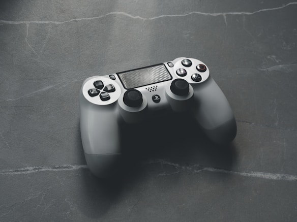A gaming controller with a sleek design placed on a textured dark surface. The controller features dual analog sticks, a central touchpad, and multiple buttons including directional and symbol-marked options. The lighting suggests a soft and cool ambiance.