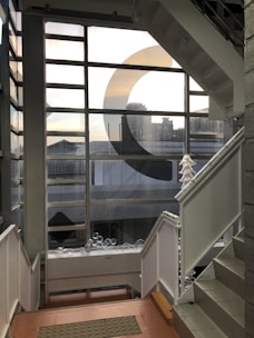 A modern stairwell with metal and glass railings, featuring a large window with a geometric design, overlooking an urban cityscape. The window lets in some natural light, casting a subtle glow on the interior. Several transparent decorative objects are placed on a ledge, adding an artistic touch.