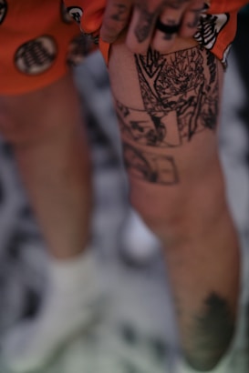 A close-up view of a person holding their thigh that features detailed black ink tattoos, with orange clothing visible in the background.