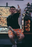 A person with tattoos sits on a desk in a room with a computer and anime figures. They wear a black long-sleeve shirt and bright orange shorts with a black and white pattern. The background features a gaming setup with a colorful, patterned screen and figurines displayed on shelves.