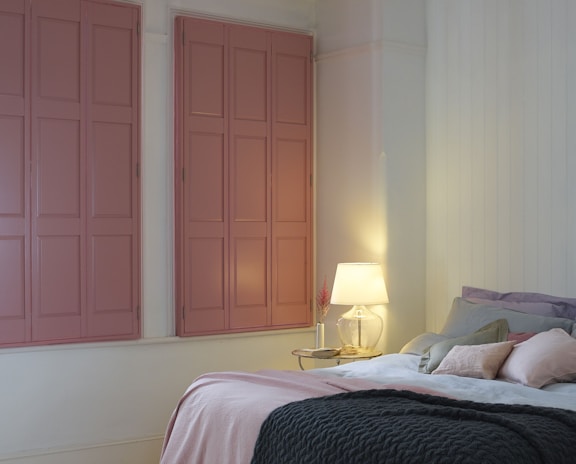 A cozy bedroom setting features a neatly made bed with pastel-colored pillows and a thick knitted blanket. Two large windows with closed, muted pink shutters provide a minimalist backdrop. A bedside table holds a glass lamp with a soft glow, enhancing the room's warm atmosphere.