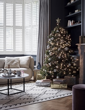 A cozy living room with a decorated Christmas tree that has lights and ornaments. The room features a modern aesthetic with large windows covered by shutters and curtains, a comfortable sofa with plush pillows, and a coffee table holding decorative items. Gift boxes sit beneath the tree, adding a festive touch.