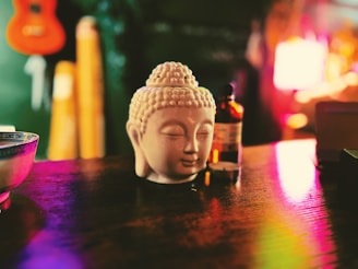 A ceramic Buddha head sits on a wooden surface next to a small bottle. The table reflects vibrant, colorful lights creating an ambient and warm atmosphere. In the background, there is a blurred view of a guitar and other objects contributing to a cozy and artistic setting.