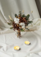 A bouquet of various dried and green foliage is artistically arranged on a soft, white fabric background. The bouquet includes cotton buds, brown hydrangea, pine branches, and small white flowers. Two lit tealight candles are placed in front of the bouquet, casting a warm glow.