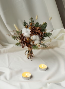 A bouquet of various dried and green foliage is artistically arranged on a soft, white fabric background. The bouquet includes cotton buds, brown hydrangea, pine branches, and small white flowers. Two lit tealight candles are placed in front of the bouquet, casting a warm glow.