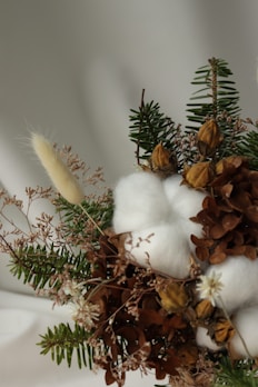 A detailed arrangement of dried flowers and plants, including green pine branches, brown leaves, fluffy white cotton, and small dried yellow buds. The natural textures and colors blend gently, suggesting a rustic and organic aesthetic.